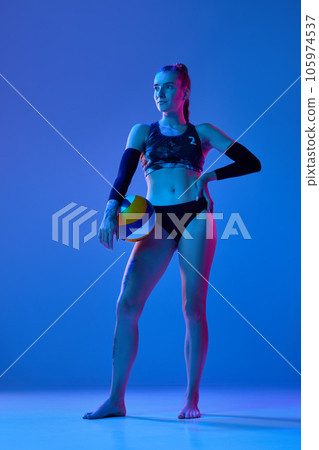 Full-length image of sportive young woman, volleyball player in sportswear standing with ball against blue studio background in neon light Full-length image of sportive young woman, volleyball player in sportswear standing with ball against blue studio background in neon light 105974537