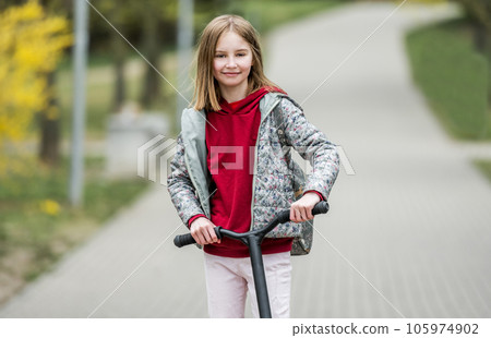 Little cute girl riding a scooter on a path in the park, smiling and looking into the camera Little cute girl riding a scooter on a path in the park, smiling and looking into the camera 105974902