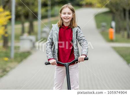 Little cute girl riding a scooter on a path in the park, smiling and looking into the camera Little cute girl riding a scooter on a path in the park, smiling and looking into the camera 105974904