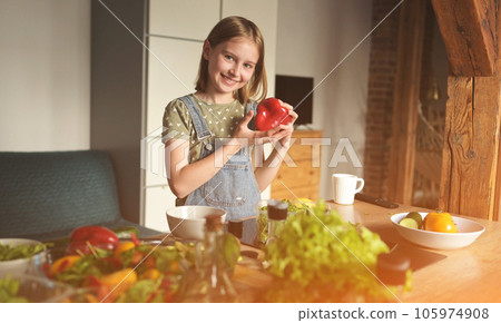 Girl child holding red sweet pepper at kitchen and smiling. Pretty female kid with paprika vegetable preparing vegetarian salad Girl child holding red sweet pepper at kitchen and smiling. Pretty female kid with paprika vegetable preparing vegetarian salad 105974908