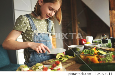 Girl child cuts avicado with knife at kitchen. Pretty female kid with fruits and vegetables preparing salad Girl child cuts avicado with knife at kitchen. Pretty female kid with fruits and vegetables preparing salad 105974911