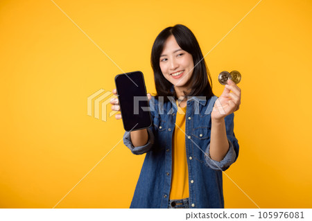 Happy young asian woman wearing yellow t-shirt denim shirt holding digital crypto currency coin and smartphone isolated on yellow background. Digital currency financial concept. Happy young asian woman wearing yellow t-shirt denim shirt holding digital crypto currency coin and smartphone isolated on yellow background. Digital currency financial concept. 105976081