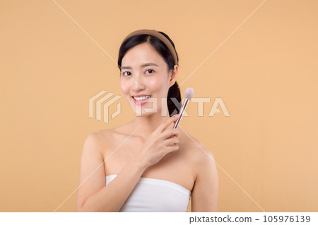 Portrait happy young asian woman with natural make up face holding cosmetic skin powder blusher isolated on beige background. Female apply skincare brush treatment. beauty product cosmetology concept. 105976139