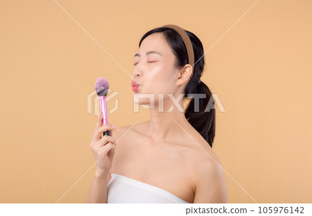 Portrait happy young asian woman with natural make up face holding cosmetic skin powder blusher isolated on beige background. Female apply skincare brush treatment. beauty product cosmetology concept. 105976142