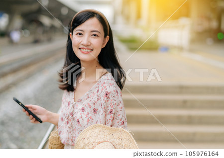 Asian young woman traveler with weaving hat using a mobile phone beside railway train station in Bangkok. Journey trip lifestyle, world travel explorer or Asia summer tourism concept. Asian young woman traveler with weaving hat using a mobile phone beside railway train station in Bangkok. Journey trip lifestyle, world travel explorer or Asia summer tourism concept. 105976164