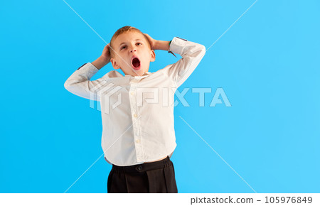 Emotional little boy, child, pupil in white shirt standing with shocked face against blue studio background 105976849