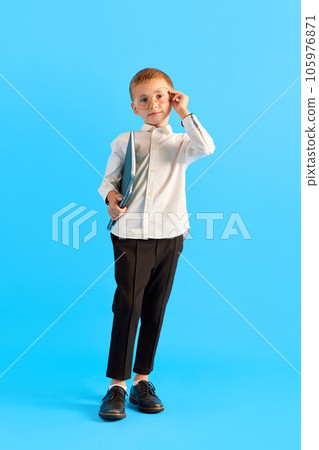 Little boy, child in glasses, wearing white shirt and black pants standing with folders against blue studio background. Smart 105976871