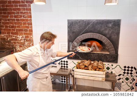 The chef prepares pizza. Raw pizza ready to bake. Cook in a apron in the kitchen. with a shovel in his hands. boxes for food delivery on background. 105981842