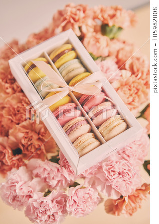 Dessert French Macarons in present box on a background of flowers. Sweet dessert on a flowers background. Still life with macaroni cakes and different flower. Delivery concept, coffee and flower shop Dessert French Macarons in present box on a background of flowers. Sweet dessert on a flowers background. Still life with macaroni cakes and different flower. Delivery concept, coffee and flower shop 105982008