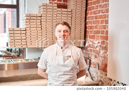 The chef prepares pizza in a neapolitan pizzeria. Cook in a apron in the kitchen with a shovel in his hands. boxes for food delivery on background. 105982048