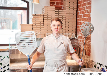 The chef prepares pizza in a neapolitan pizzeria. Cook in a apron in the kitchen with a shovel in his hands. boxes for food delivery on background. 105982049
