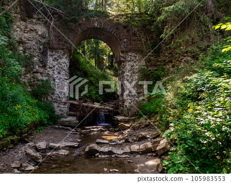 Old stone arched destroyed bridge over small river in a mountain forest 105983553