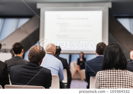 Business and entrepreneurship symposium. Female speaker giving a talk at business meeting. Audience in conference hall. Rear view of unrecognized participant in audience. 105984582