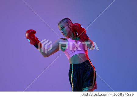 Sportive teen girl, MMA athlete in uniform and boxing gloves, training against purple studio background in neon lights Sportive teen girl, MMA athlete in uniform and boxing gloves, training against purple studio background in neon lights 105984589
