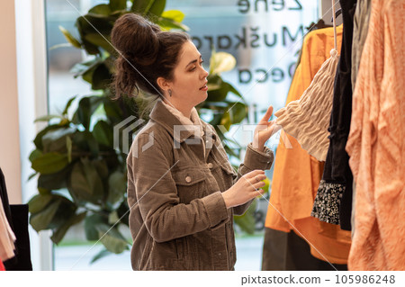 Side view of young Caucasian pretty woman choices dress in shop. Shopping and sale Side view of young Caucasian pretty woman choices dress in shop. Shopping and sale 105986248