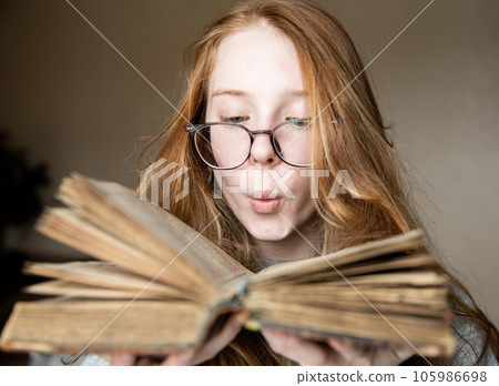 Red-haired beautiful girl with glasses blowing dust off a vintage old book. 105986698