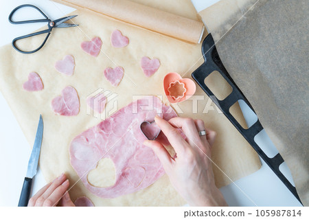 The chef cuts a heart-shaped sausage-ham cut into a heart-shaped shape on parchment, top view, on a pizza deco background. Cooking pizza for Valentine's Day. 105987814