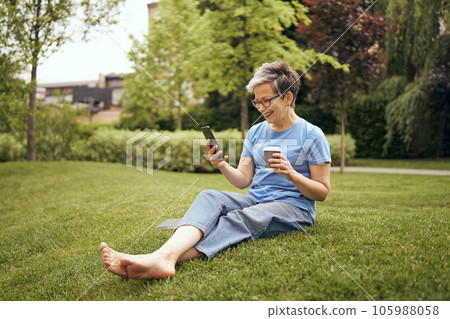 Mature woman having rest in city park, sitting on grass and drinking coffee while use phone 105988058