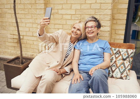Mature smiling women friends taking selfie on phone while sitting in cafe terrace Mature smiling women friends taking selfie on phone while sitting in cafe terrace 105988060
