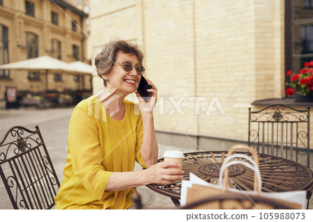 Smiling senior woman talking on smartphone while holding coffee cup at cafe terrace after shopping Smiling senior woman talking on smartphone while holding coffee cup at cafe terrace after shopping 105988073