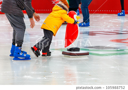 A child, accompanied by an adult, learns to skate on an ice rink, holding on to an assistant, a toy penguin. A child, accompanied by an adult, learns to skate on an ice rink, holding on to an assistant, a toy penguin. 105988752