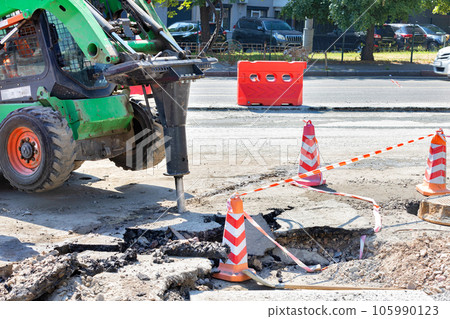 The barrel of a powerful jackhammer on a maneuverable and nimble road tractor breaks the old asphalt on a summer day. The barrel of a powerful jackhammer on a maneuverable and nimble road tractor breaks the old asphalt on a summer day. 105990123