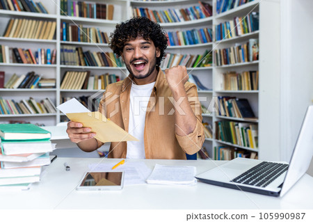 Portrait of young successful student, man received letter of notification of good results of interview exam, hispanic man joyfully looking at camera inside library, holding mail envelope. 105990987