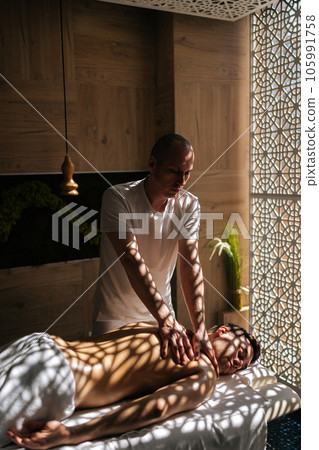 Portrait of professional masseur male with strong hands doing sports massage to back muscles of sportsman lying on massage table by window. Physiotherapist making wellness massage to athlete client. 105991758