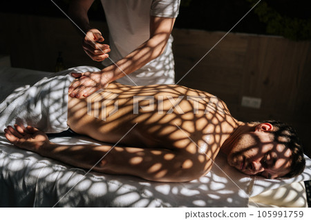 Cropped shot of unrecognizable manual therapist massaging man back with oil during relaxing or revitalizing procedure at Thai beauty spa salon. Guy client lying on couch enjoying body and skin care. 105991759