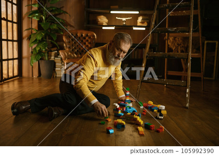 Senior old man playing toy building blocks while sitting on home floor 105992390