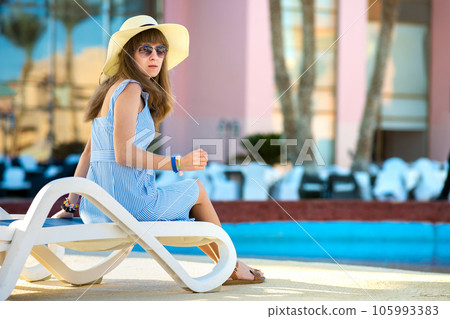 Young woman wearing light summer dress and yellow straw hat sitting outside near hotel swimming pool on summer sunny day. 105993383