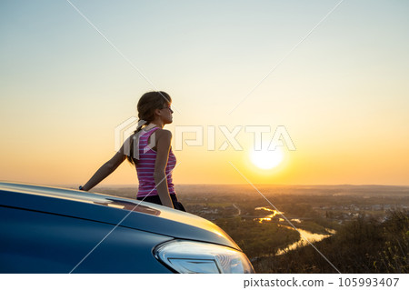 Young woman driver standing near her car enjoying warm sunset view. Girl traveler leaning on vehicle hood looking at evening horizon. Young woman driver standing near her car enjoying warm sunset view. Girl traveler leaning on vehicle hood looking at evening horizon. 105993407