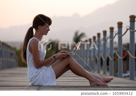 Young woman browsing internet on laptop computer outdoors on warm summer evening. Remote work and study concept. Young woman browsing internet on laptop computer outdoors on warm summer evening. Remote work and study concept. 105993412