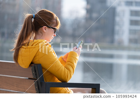 Young pretty woman sitting on a park bench browsing her smartphone outdoors on warm autumn day. Young pretty woman sitting on a park bench browsing her smartphone outdoors on warm autumn day. 105993424