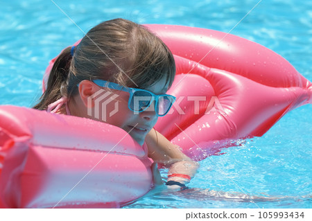 Young joyful child girl having fun swimming on inflatable air mattress in swimming pool with blue water on warm summer day on tropical vacations. Summertime activities concept Young joyful child girl having fun swimming on inflatable air mattress in swimming pool with blue water on warm summer day on tropical vacations. Summertime activities concept 105993434