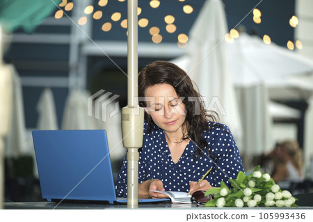 Young happy woman working remotely on laptop computer sitting at outdoor restaurant table. Doing business online concept 105993436