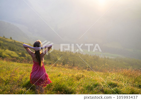Young happy woman traveler in red dress standing on grassy hillside on a windy evening in summer mountains enjoying view of nature at sunset. 105993437