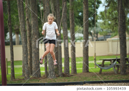 Young happy child girl playing alone, flying high on swings on summer weekend sunny day. Safety and recreation on playground concept Young happy child girl playing alone, flying high on swings on summer weekend sunny day. Safety and recreation on playground concept 105993439