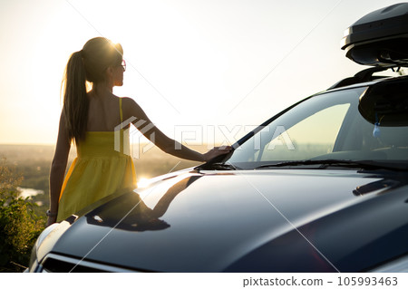 Yong happy female driver resting near her car enjoying sunset view of summer nature. Travel and recreation concept. 105993463