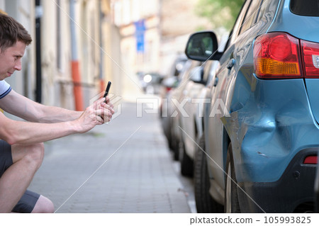 Stressed driver taking picture on sellphone camera of smashed vehicle parked on street side calling for emergency service help after car accident. Road safety and insurance concept 105993825