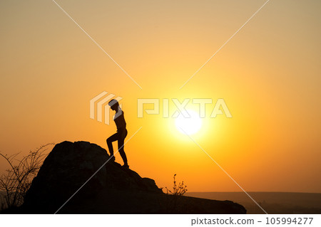 Silhouette of a woman hiker climbing up a big stone at sunset in mountains. Female tourist on high rock in evening nature. Tourism, traveling and healthy lifestyle concept. 105994277