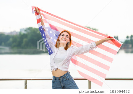 Portrait of happy smiling red haired girl holding USA national flag up in her hands. Positive young woman celebrating United States independence day. Portrait of happy smiling red haired girl holding USA national flag up in her hands. Positive young woman celebrating United States independence day. 105994432