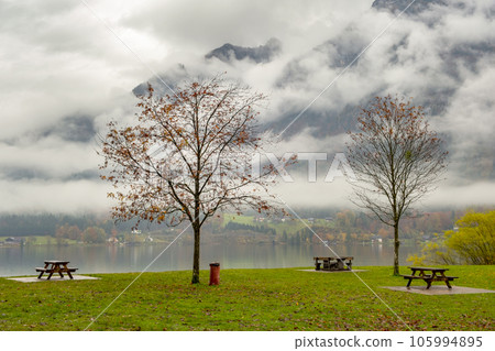 Moody autumn landscape with bare trees and benches on mountain lake shore. Moody autumn landscape with bare trees and benches on mountain lake shore. 105994895
