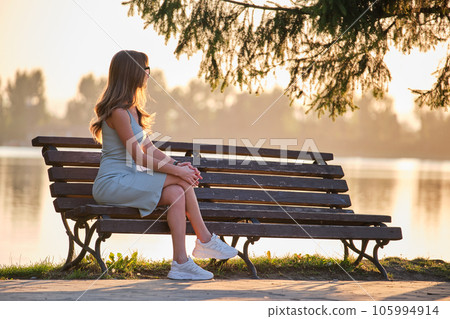 Lonely woman sitting alone on lake shore bench on warm summer evening. Solitude and relaxing in nature concept. 105994914