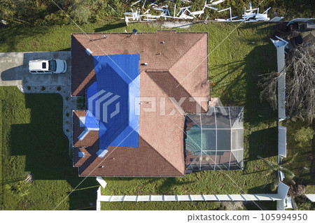 Hurricane Ian damaged house rooftop covered with protective plastic tarp against rain water leaking until replacement of asphalt shingles. Aftermath of natural disaster 105994950