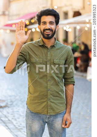 Young man smiling friendly at camera, waving hands hello, hi, greeting or goodbye in city street 105995323