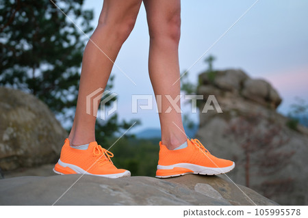 Closeup of female young slim legs in bright orange sneaker shoes standing on mountain hiking trail in summer evening. Active lifestyle, jogging in nature and exercise on fresh air concept. 105995578