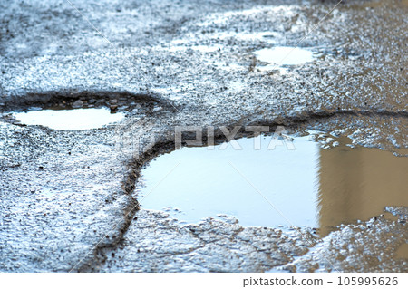 Close up of a road in very bad condition with big potholes full of dirty rain water pools. 105995626