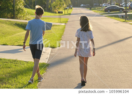 Back view of two young teenage children, girl and boy, brother and sister walking together on suburban street on bright sunny evening 105996104