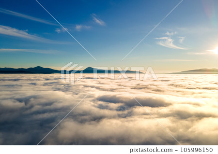 Aerial view of vibrant sunset over white dense clouds with distant dark mountains on horizon. 105996150